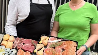 Ein Mann und eine Frau halten zwei Tabletts mit verschiedenen Fleisch- und Meeresfrüchten und lächeln vor einem Metallzaun.