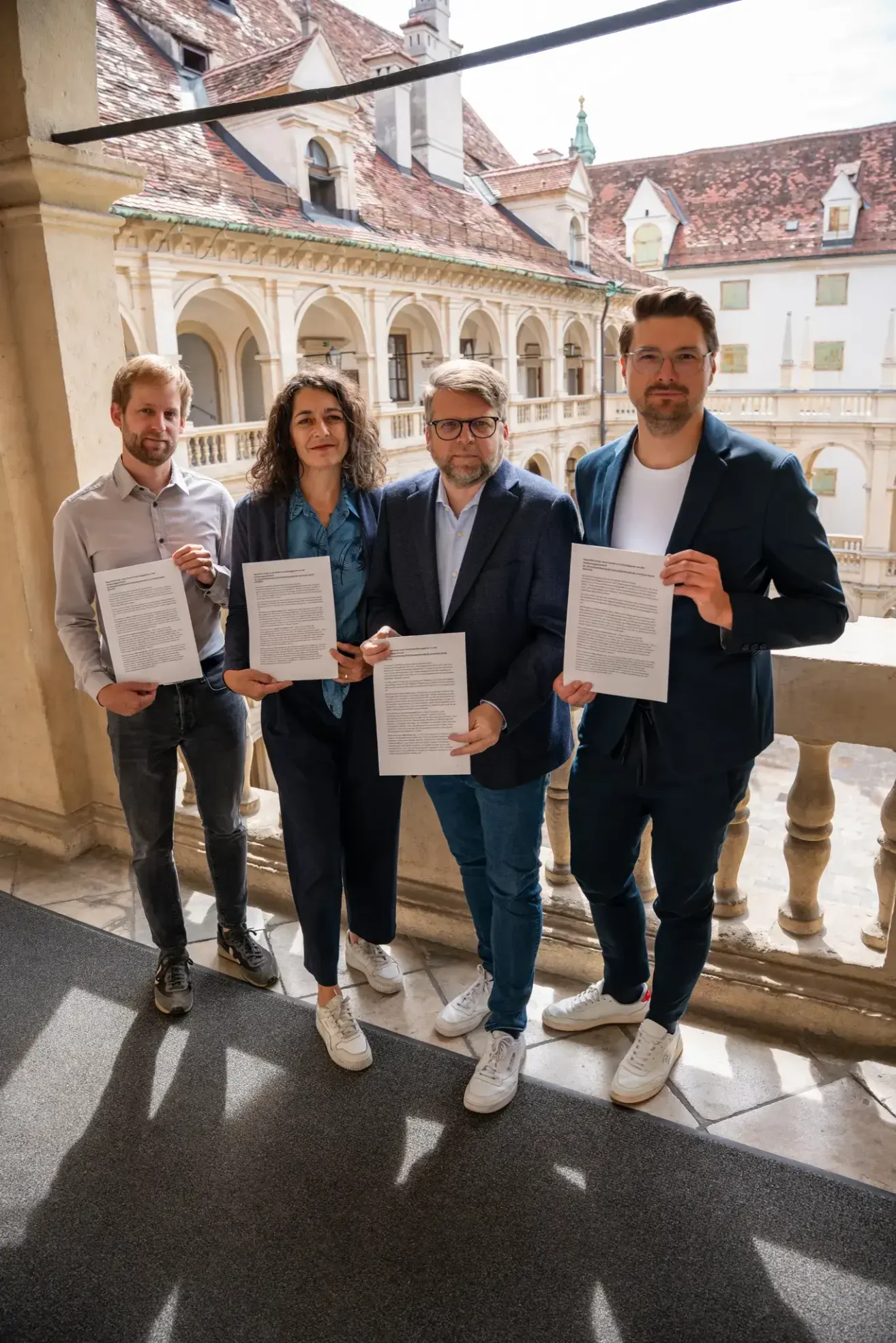 Vier Erwachsene stehen vor einem historischen Gebäude. Sie halten Papiere in den Händen und tragen formelle Kleidung. Der Mann links trägt eine Brille und ein langärmeliges Hemd.