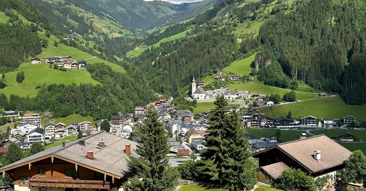 Luftaufnahme eines Bergdorfes mit dichten grünen Hügeln und einer großen weißen Kirche. Zahlreiche Häuser sind zwischen Bäumen verstreut, einige mit Balkonen.