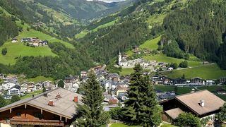 Luftaufnahme eines Bergdorfes mit dichten grünen Hügeln und einer großen weißen Kirche. Zahlreiche Häuser sind zwischen Bäumen verstreut, einige mit Balkonen.