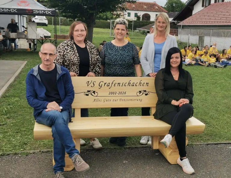 Fünf Menschen sitzen auf einer Bank im Freien. Ein Mann in einer blauen Jacke und einer Brille sitzt links. Eine Frau sitzt rechts. Die Bank hat eine Holztafel mit der Aufschrift VS Grafenschachen 2002-2026.