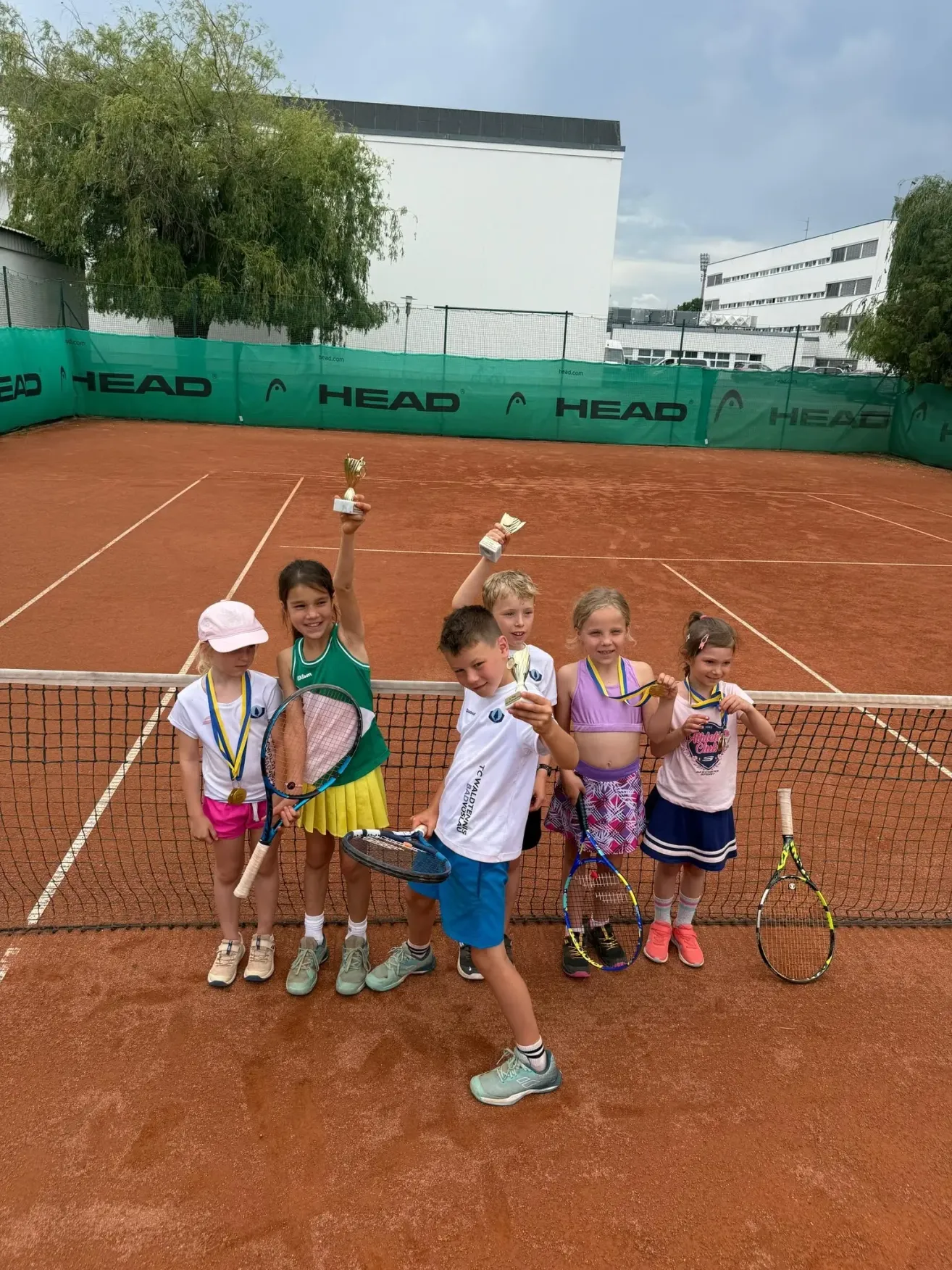 Eine Gruppe junger Tennisspieler, alle mit Pokalen, steht auf einem Tennisplatz und posiert lächelnd für ein Foto.