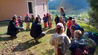 Eine Gruppe von Kindern und Erwachsenen steht draußen in einem Grasbereich. Einige tragen Mützen, Turnschuhe und haben Rucksäcke dabei. Sie sind in der Nähe eines Gebäudes mit Fenstern und Bergen in der Ferne.