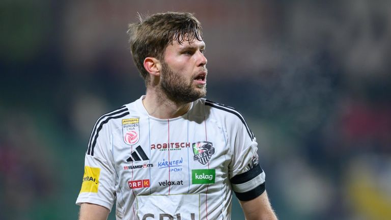 Ein Mann in einem Fußballtrikot mit verschiedenen Sponsorenlogos, darunter Adidas, steht und scheint zu sprechen.