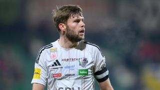 Ein Mann in einem Fußballtrikot mit verschiedenen Sponsorenlogos, darunter Adidas, steht und scheint zu sprechen.