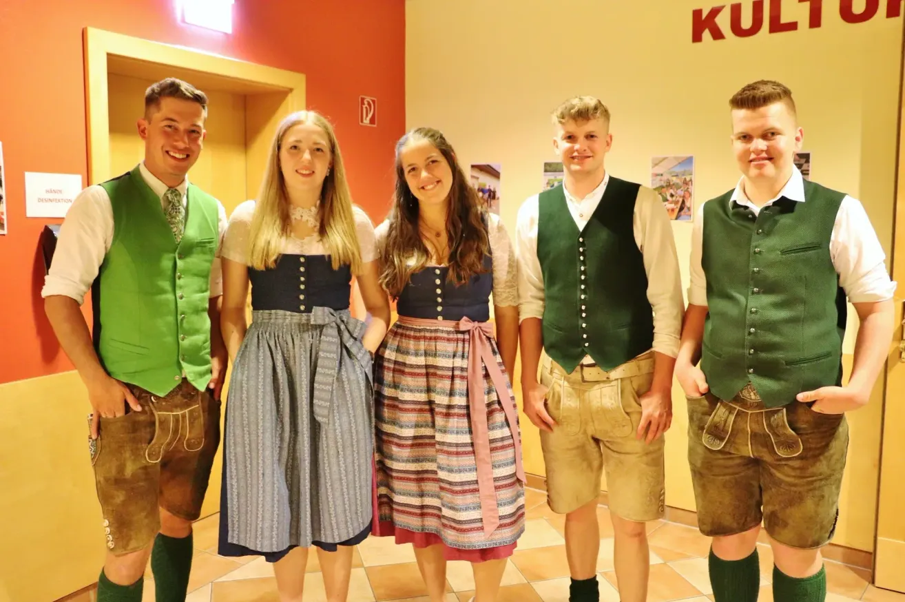 Fünf Personen stehen in einem Raum und sind in traditioneller Kleidung gekleidet. Die beiden Frauen tragen Dirndl und die drei Männer tragen Westen und Shorts. Sie lächeln für die Kamera.