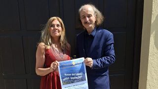 Ein Mann und eine Frau posieren für ein Foto, der Mann hält ein Plakat mit dem Titel 'Wasser des Lebens' auf Deutsch, was auf eine Veranstaltung oder Zusammenkunft hindeutet.