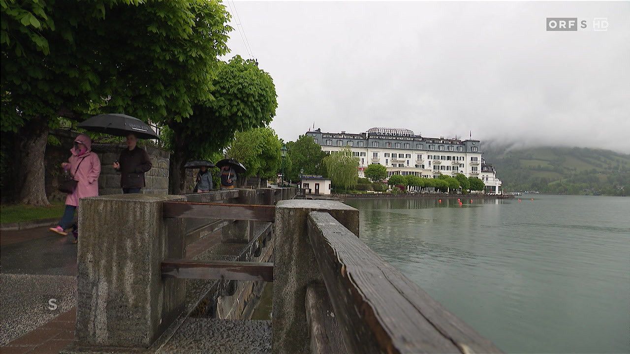 Blick auf einen See mit einem großen Hotel im Hintergrund. Zwei Personen mit Schirmen gehen links. Das Wetter ist bewölkt.