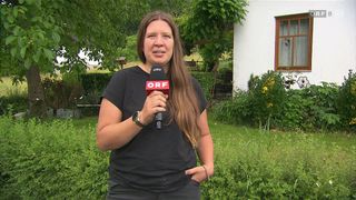 Eine Frau steht im Garten und hält ein Mikrofon mit roten und weißen Buchstaben. Sie trägt ein schwarzes Shirt und Hose. Hinter ihr sind Büsche, Bäume und ein weißes Haus mit Fenster.