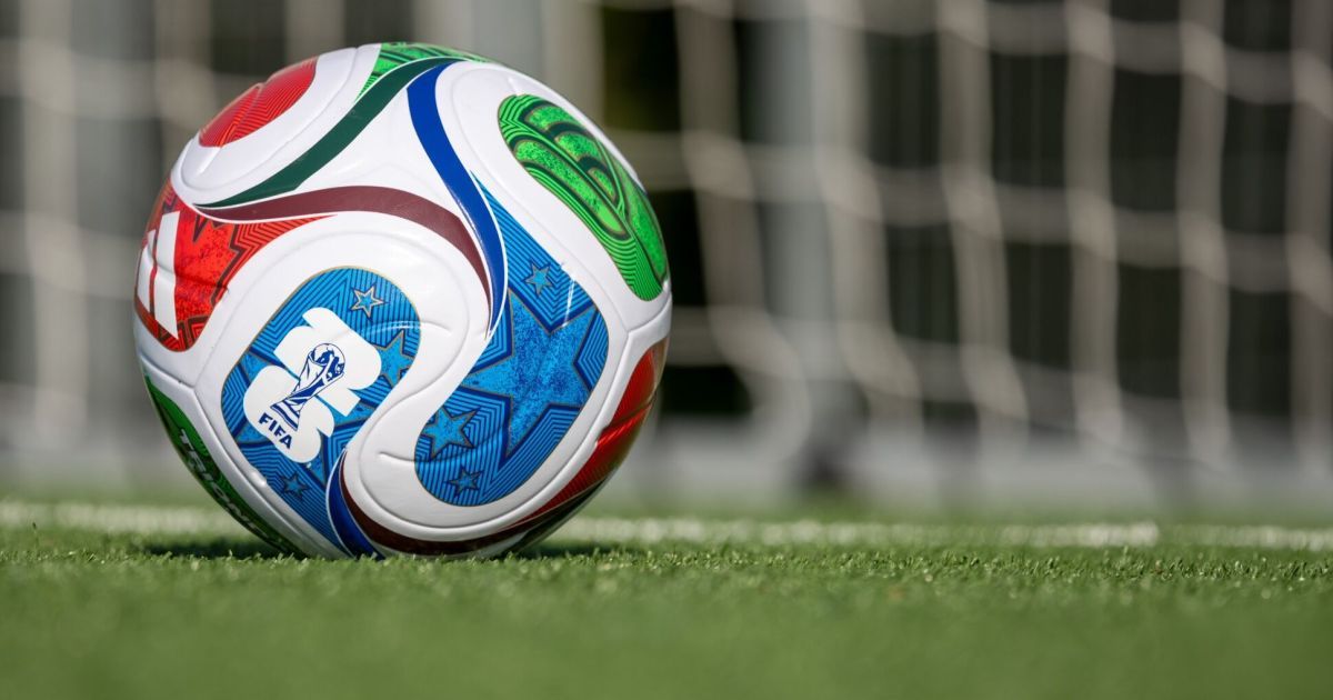 Ein farbenfroher Fußball mit dem FIFA-WM-Logo liegt auf einem grünen Feld. Der Hintergrund zeigt eine verschwommene Ansicht eines Fußballtors.