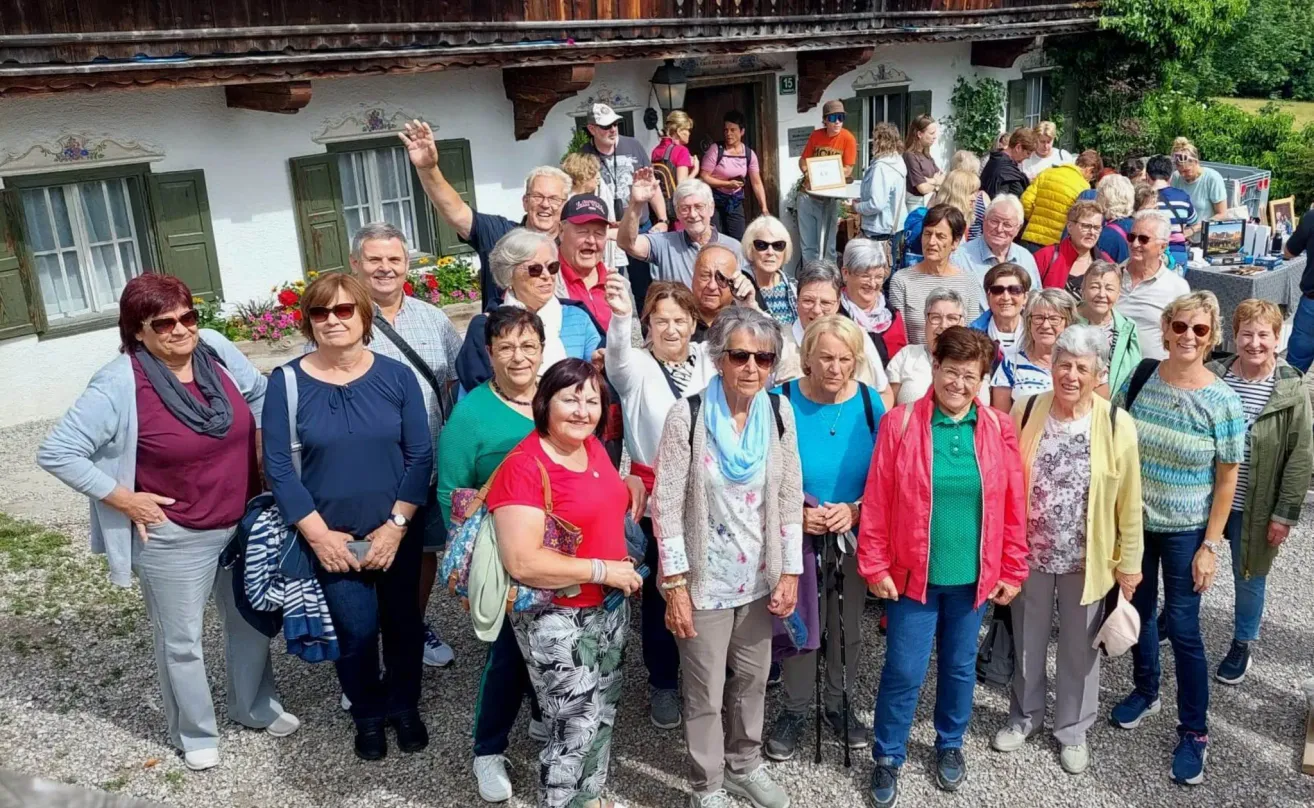 Eine Gruppe älterer Menschen posiert für ein Foto vor einem Chalet. Einige lächeln, andere tragen Sonnenbrillen. Dahinter ist ein Gebäude mit grünen Fensterläden und einer Laterne zu sehen.