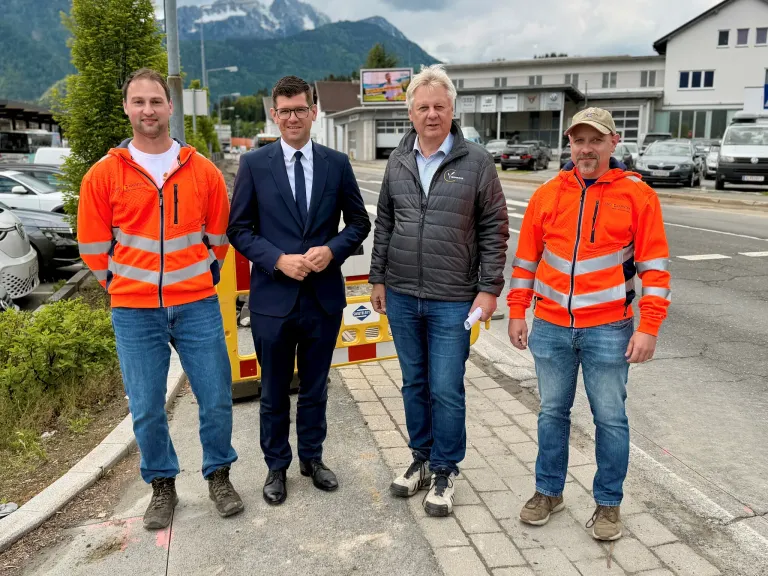 Vier Männer stehen auf einem Bürgersteig mit Bergen im Hintergrund. Einer hält ein Papier. Sie tragen orange Sicherheitsjacken und Jeans.