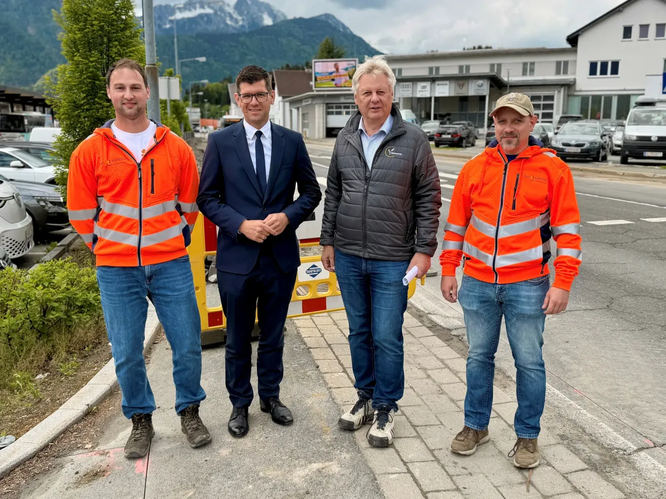 Vier Männer stehen auf einem Bürgersteig mit Bergen im Hintergrund. Einer hält ein Papier. Sie tragen orange Sicherheitsjacken und Jeans.