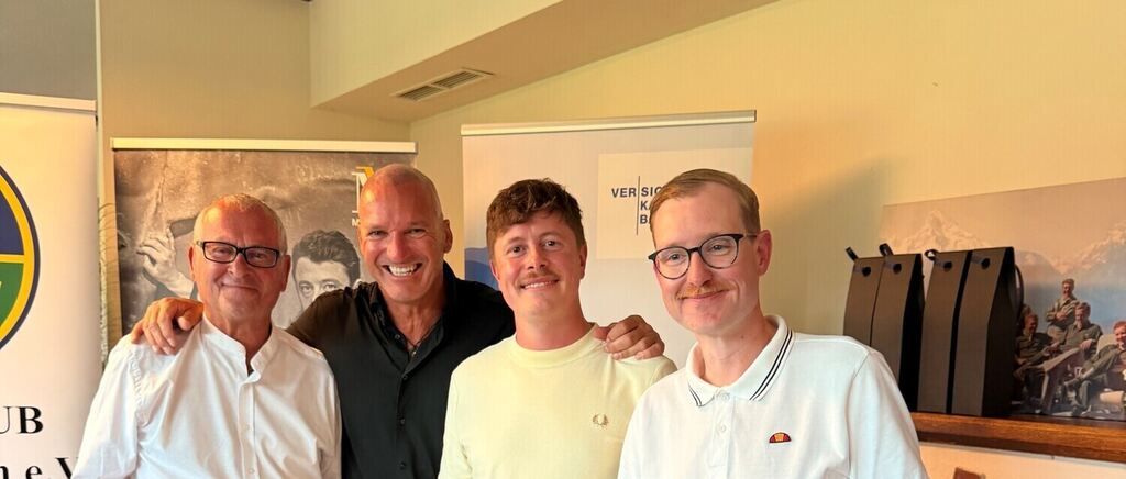 Four men stand smiling in a room. One man has his arm around another. They wear casual clothing. A whiteboard and a photo are in the background.