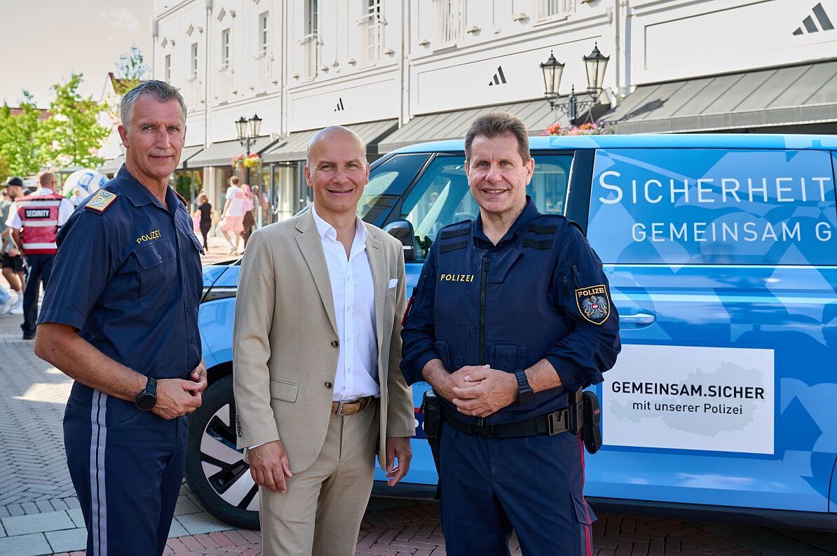 Drei Männer stehen vor einem blauen Polizeivan. Zwei tragen Polizeiuniformen, einer trägt einen beigen Anzug. Sie alle lächeln für ein Foto.