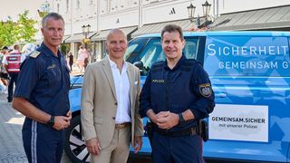 Drei Männer stehen vor einem blauen Polizeivan. Zwei tragen Polizeiuniformen, einer trägt einen beigen Anzug. Sie alle lächeln für ein Foto.