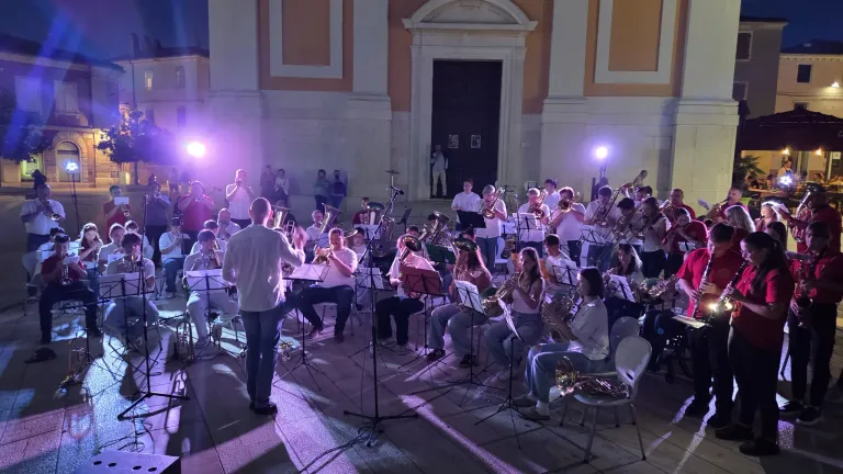 Ein Orchester spielt in einem Außenbereich mit lila Beleuchtung. Einige sitzen und einige stehen und spielen Musikinstrumente.