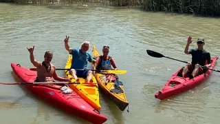 Drei Menschen paddeln auf einem ruhigen Fluss. Ein Mann gibt ein Friedenszeichen. Zwei Kajaks sind gelb, eines rot.