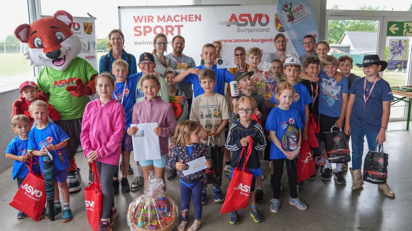 Eine Gruppe von Kindern und Erwachsenen steht gemeinsam vor einem Banner mit den Texten 'Wir machen Sport' und 'ASVO'. Sie halten Tassen, Taschen und Geschenke. Einige von ihnen tragen Brillen und Sneakers.