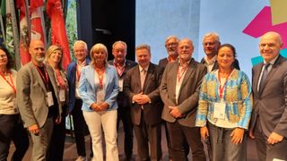 Eine Gruppe von Menschen in Anzügen und Krawatten steht vor einer Wand mit einem Baum und einer Flagge. Sie nehmen an einer Veranstaltung teil.