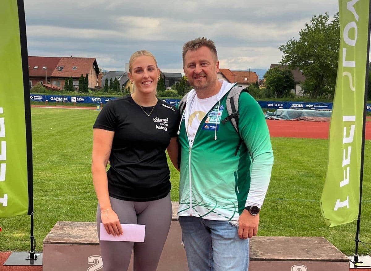 Ein Mann und eine Frau stehen auf einem Sportplatz, lächeln und posieren für ein Foto. Die Frau hält ein Stück Papier in der Hand. Hinter ihnen befindet sich ein Rasenfeld mit einer Laufbahn und Häusern.