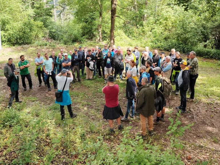 Eine Gruppe von Menschen steht im Kreis in einem bewaldeten Gebiet, einige reden und andere lauschen. Es gibt mehrere Bäume und Pflanzen in der Umgebung.