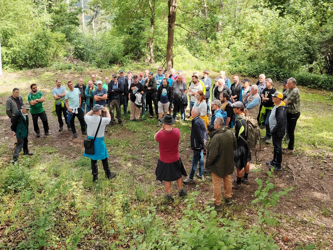 Eine Gruppe von Menschen steht im Kreis in einem bewaldeten Gebiet, einige reden und andere lauschen. Es gibt mehrere Bäume und Pflanzen in der Umgebung.