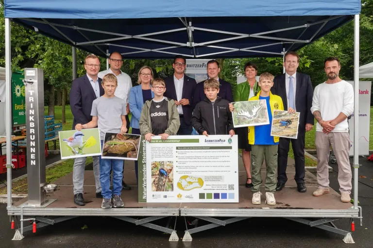 Eine Gruppe von Erwachsenen und Kindern steht unter einem Zelt, hält Plakate und ein Schild in der Hand. Die Kinder lächeln und die Erwachsenen sind formell gekleidet. Sie sind wahrscheinlich bei einer Veranstaltung.