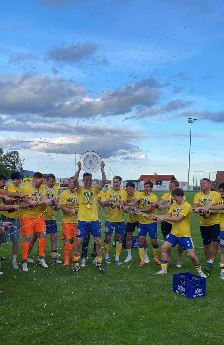 Fußballspieler feiern auf einem Feld und halten einen Pokal. Sie tragen gelbe Trikots, Shorts und Turnschuhe. Ein Spieler hält einen weißen Teller, während andere Getränke einschenken. Im Hintergrund sind eine bewölkte Himmelslandschaft, Bäume und eine Straßenlaterne zu sehen.