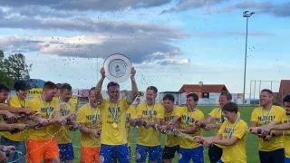 Fußballspieler feiern auf einem Feld und halten einen Pokal. Sie tragen gelbe Trikots, Shorts und Turnschuhe. Ein Spieler hält einen weißen Teller, während andere Getränke einschenken. Im Hintergrund sind eine bewölkte Himmelslandschaft, Bäume und eine Straßenlaterne zu sehen.