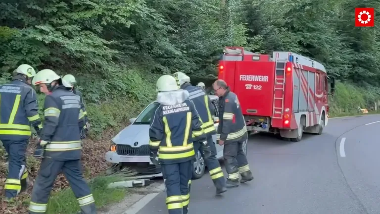 Feuerwehrleute überprüfen ein beschädigtes Fahrzeug am Straßenrand, ein roter Feuerwehrwagen ist in der Nähe.