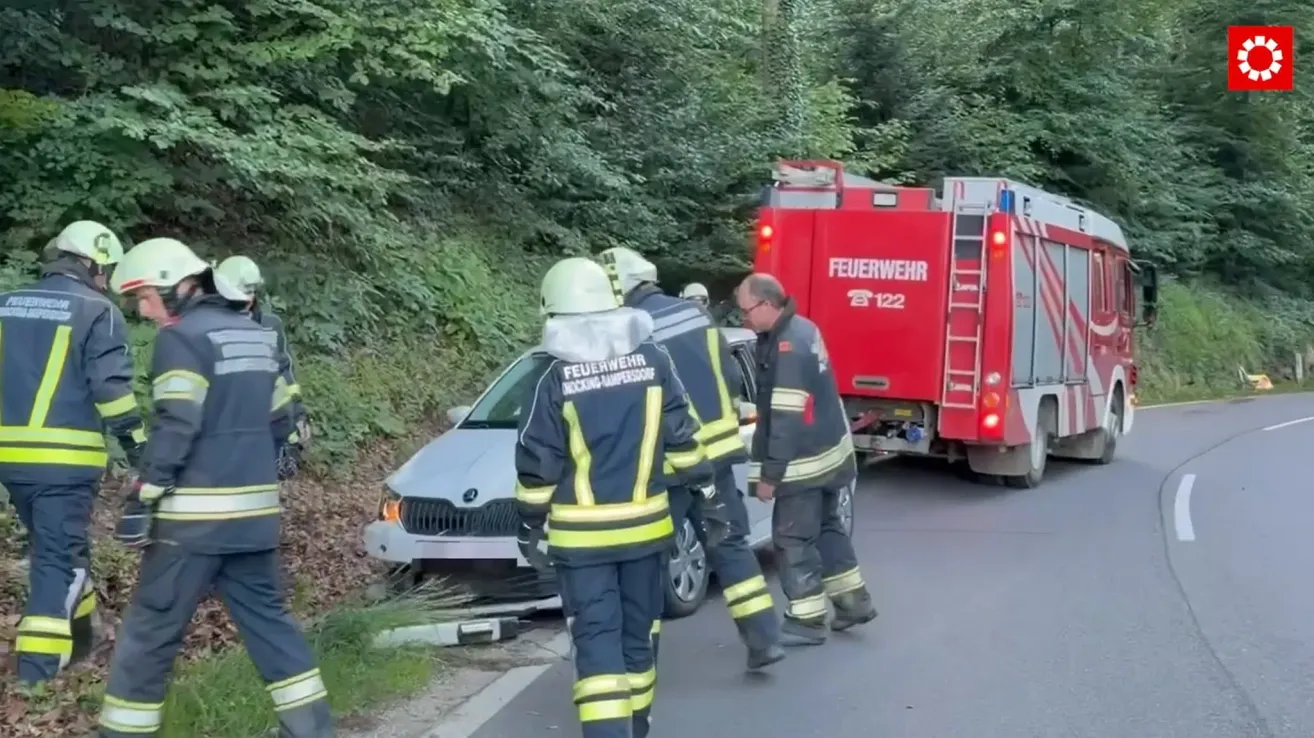 Feuerwehrleute überprüfen ein beschädigtes Fahrzeug am Straßenrand, ein roter Feuerwehrwagen ist in der Nähe.