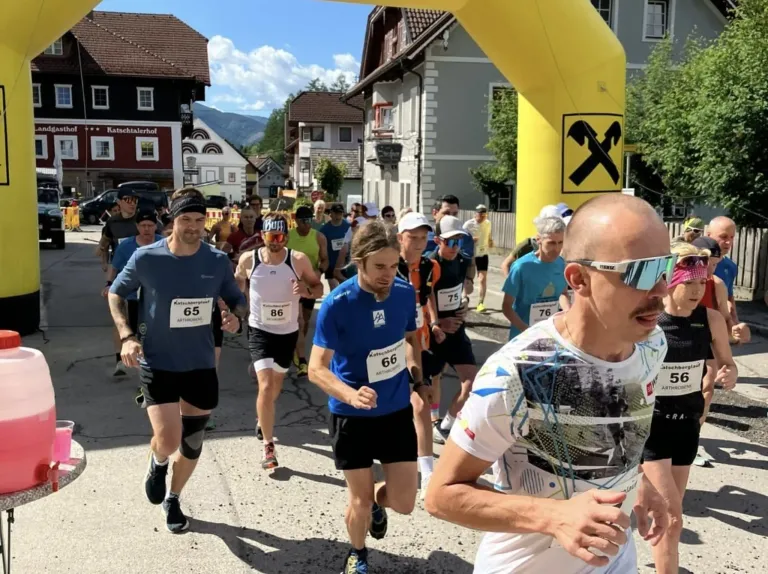 Teilnehmer laufen in einem Marathon mit Gebäuden und Bergen im Hintergrund. Die Läufer haben Nummern auf ihren Trikots.