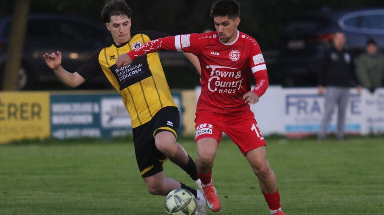 Zwei Fußballspieler sind auf einem Feld. Einer trägt ein gelbes und schwarzes Trikot, der andere ein rotes Trikot mit 'Town & Country Haus' darauf. Sie konkurrieren um den Ball.