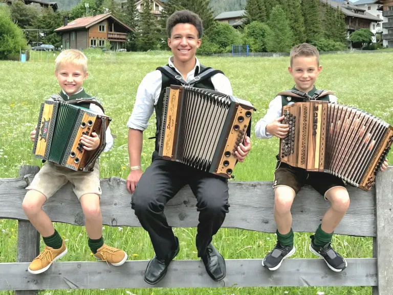 Drei Jungen in traditioneller Kleidung sitzen auf einem Holzzaun und halten Akkordeons. Der mittlere Junge lächelt, während die anderen nach vorne schauen. Dahinter sind ein Grasfeld und Häuser zu sehen.