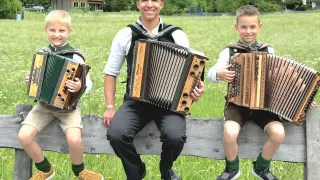 Drei Jungen in traditioneller Kleidung sitzen auf einem Holzzaun und halten Akkordeons. Der mittlere Junge lächelt, während die anderen nach vorne schauen. Dahinter sind ein Grasfeld und Häuser zu sehen.