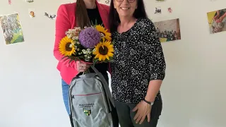 Zwei Frauen stehen zusammen in einem Spielzimmer, eine hält einen Strauß Sonnenblumen und einen Rucksack, die andere lächelt. Hinter ihnen ist eine weiße Wand mit Kinderzeichnungen und dem Wort 'Baby Reff'.