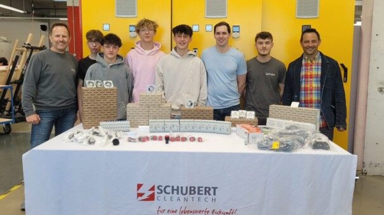 Sieben junge Männer stehen hinter einem Tisch mit verschiedenen Gegenständen darauf. Sie stehen vor einer gelben Wand mit einem Logo, das Schubert Cleantech liest.