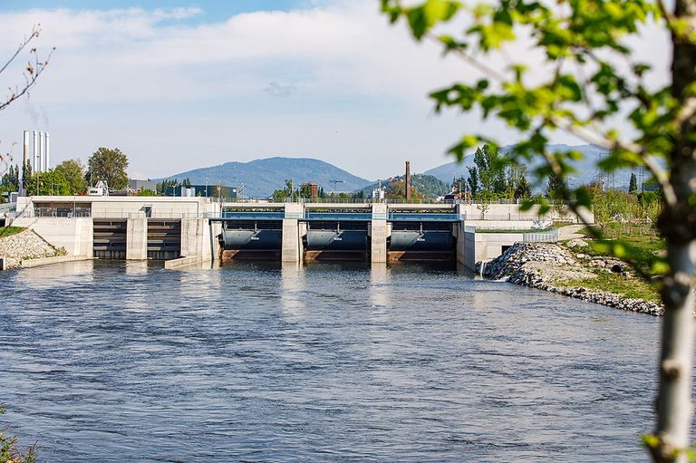Ein großer Damm mit mehreren Schleusentoren befindet sich entlang eines Flusses mit Bergen im Hintergrund.