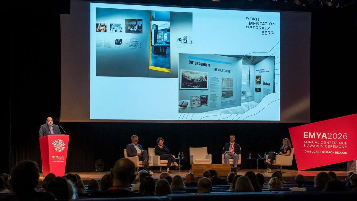 A presentation on stage with a projector screen behind it, showing images and text. Four people sit in chairs, facing the audience. The audience sits in front of them.
