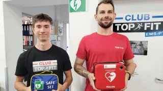 Zwei Männer stehen nebeneinander vor einer Wand. Der Mann auf der linken Seite hält eine blaue und gelbe Tasche mit der Aufschrift 'ARKY SAFE SET'. Der Mann auf der rechten Seite hält einen roten Philips-Defibrillator. Hinter ihnen befindet sich ein grün-weißes Schild mit einem Blitz und einem Herzsymbol.