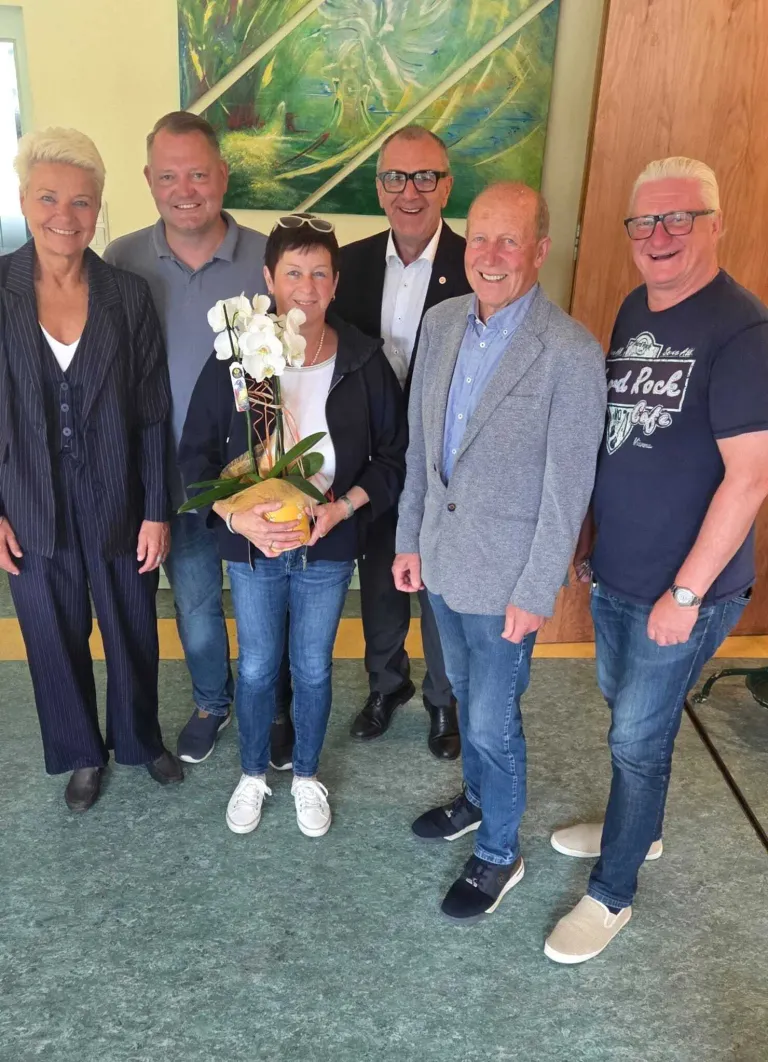 Eine Gruppe von Menschen steht drinnen und lächelt für ein Foto. Eine Frau hält einen Strauß weißer Blumen. Hinter ihnen ist eine Wand mit einem Gemälde.