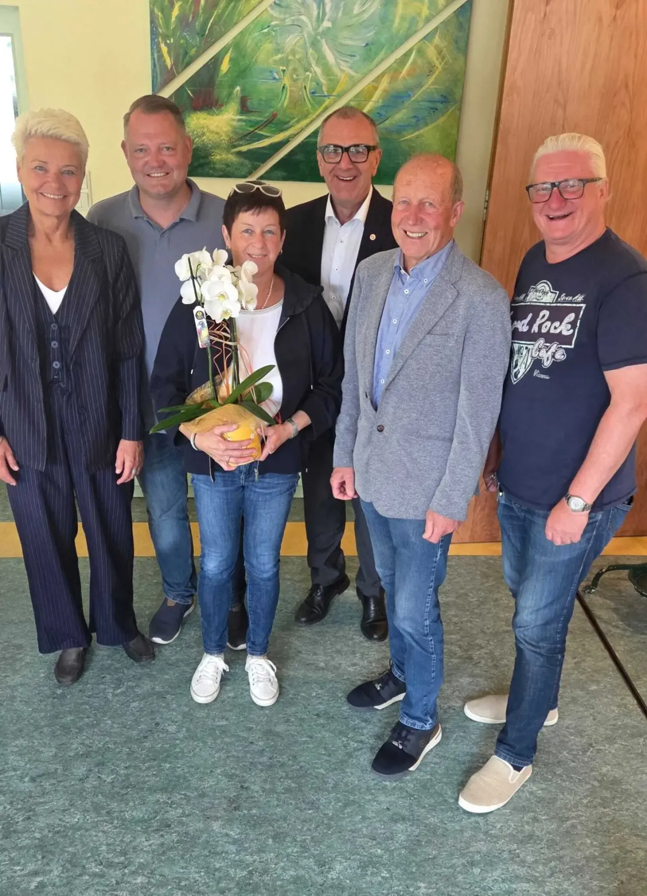 Eine Gruppe von Menschen steht drinnen und lächelt für ein Foto. Eine Frau hält einen Strauß weißer Blumen. Hinter ihnen ist eine Wand mit einem Gemälde.