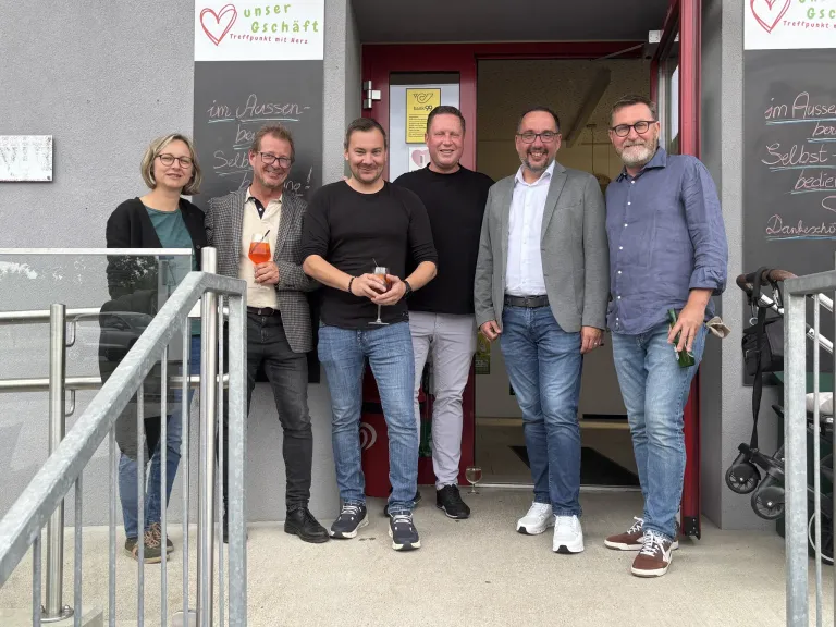 Fünf Erwachsene posieren vor einem Gebäude mit einem Schild, das Unser Geschaft steht. Sie lächeln und halten Getränke.