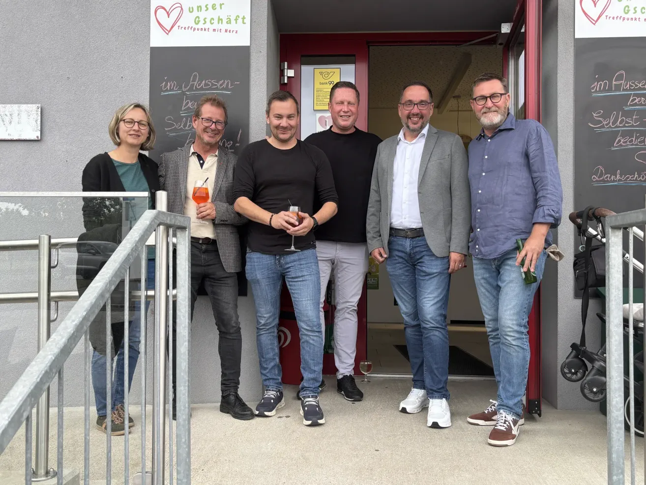 Fünf Erwachsene posieren vor einem Gebäude mit einem Schild, das Unser Geschaft steht. Sie lächeln und halten Getränke.
