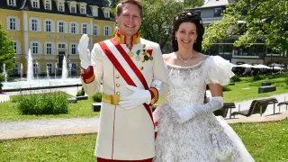 A man and woman dressed in royal attire stand in front of a fountain and a yellow building. The man wears medals, and the woman has a white dress and necklace.