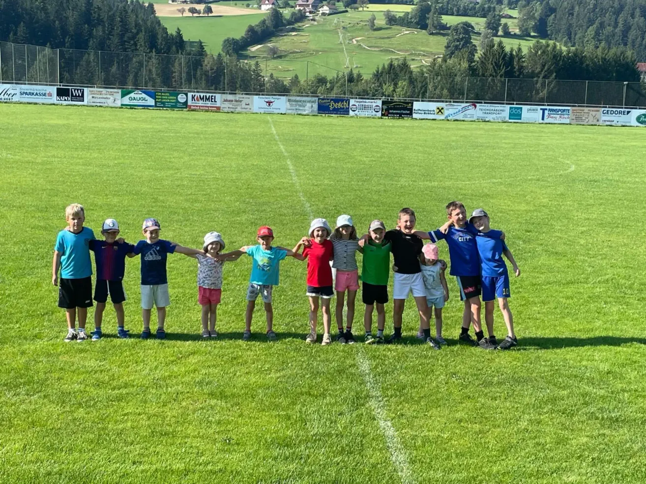 Gruppe von Kindern, die in einer Reihe auf einem Grasfeld mit einem Zaun im Hintergrund stehen. Sie alle lächeln für ein Foto.