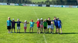 Gruppe von Kindern, die in einer Reihe auf einem Grasfeld mit einem Zaun im Hintergrund stehen. Sie alle lächeln für ein Foto.