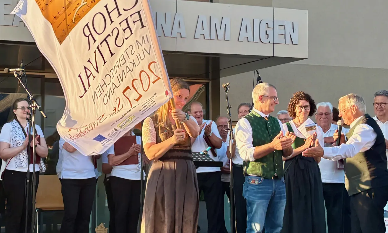 A woman holds a banner and claps at an outdoor event. Behind her, a building with a sign 'NA AM AIGEN'. An elderly man smiles at her.