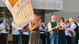 Eine Frau hält ein Banner und klatscht bei einer Veranstaltung im Freien. Dahinter befindet sich ein Gebäude mit dem Schild 'NA AM AIGEN'. Ein älterer Mann lächelt sie an.